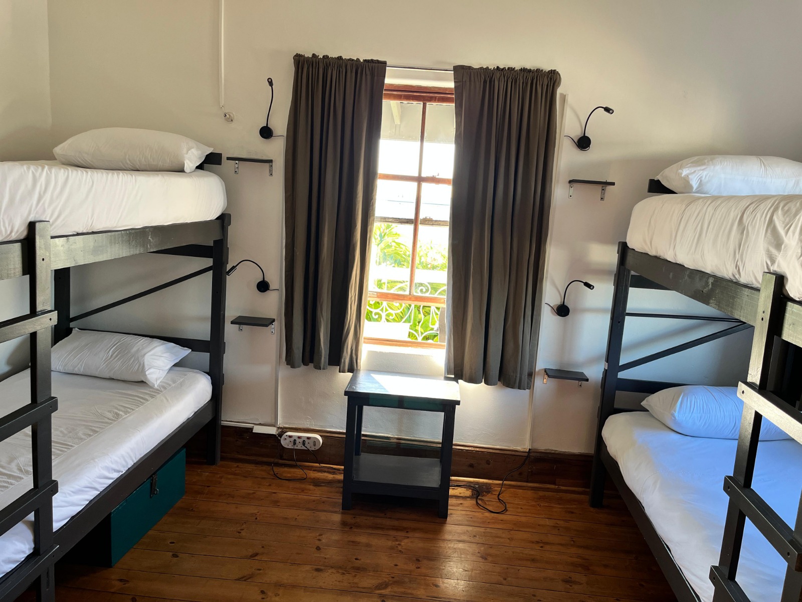 Dormitory with four black bunk beds, white bedding, wooden floor, gray curtains, tropical view