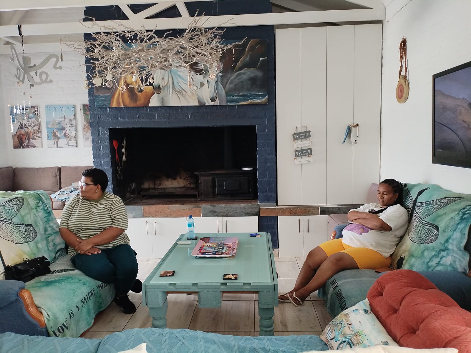 Modern living room with two people on green couches facing a light blue coffee table, dark fireplace, and coastal artwork