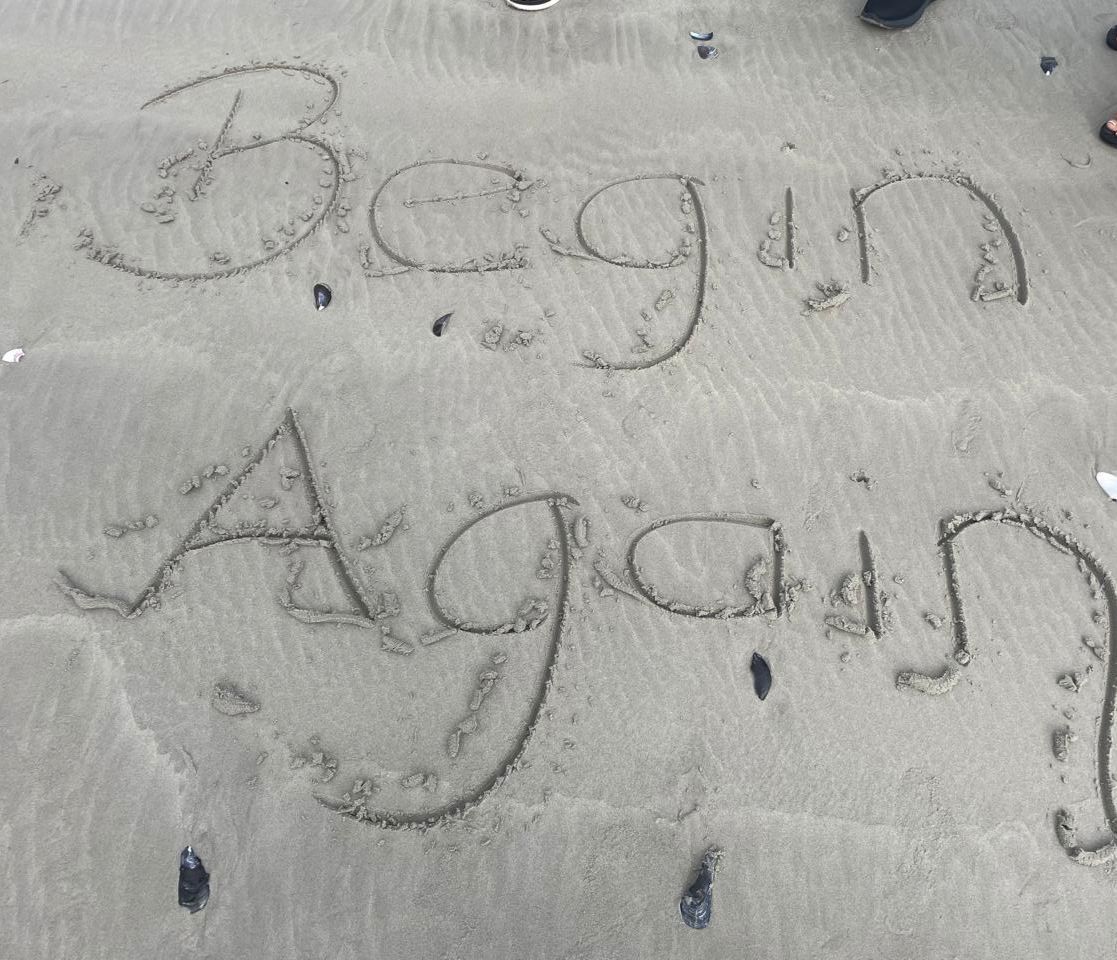 The words 'Begin Again' written in sand on a beach with seashells and small stones scattered around