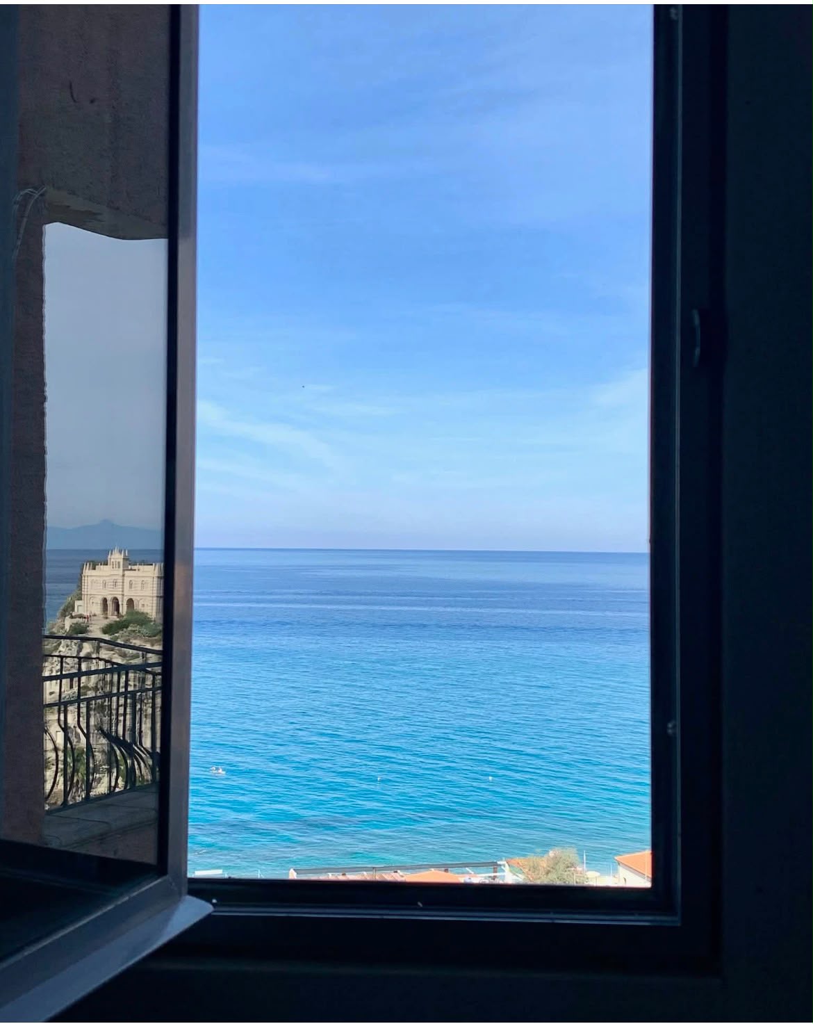 View through an open window framing a breathtaking Mediterranean coastal scene with turquoise sea, a historic castle perched on a cliff, and distant mountains under a clear blue sky