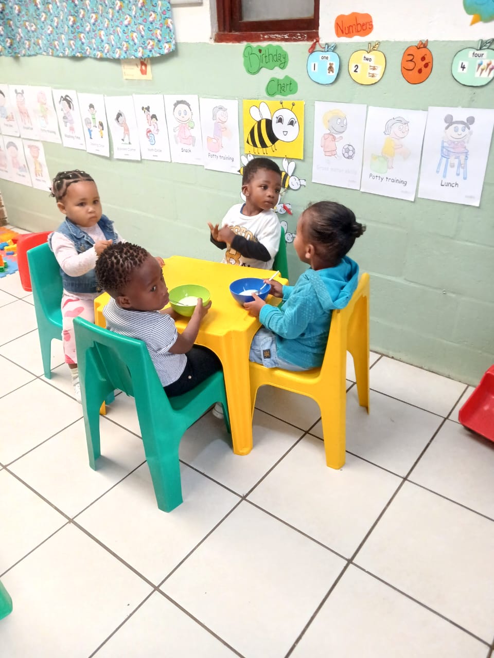Young children sitting at a colourful yellow and green table enjoying snacks together in a cheerful classroom