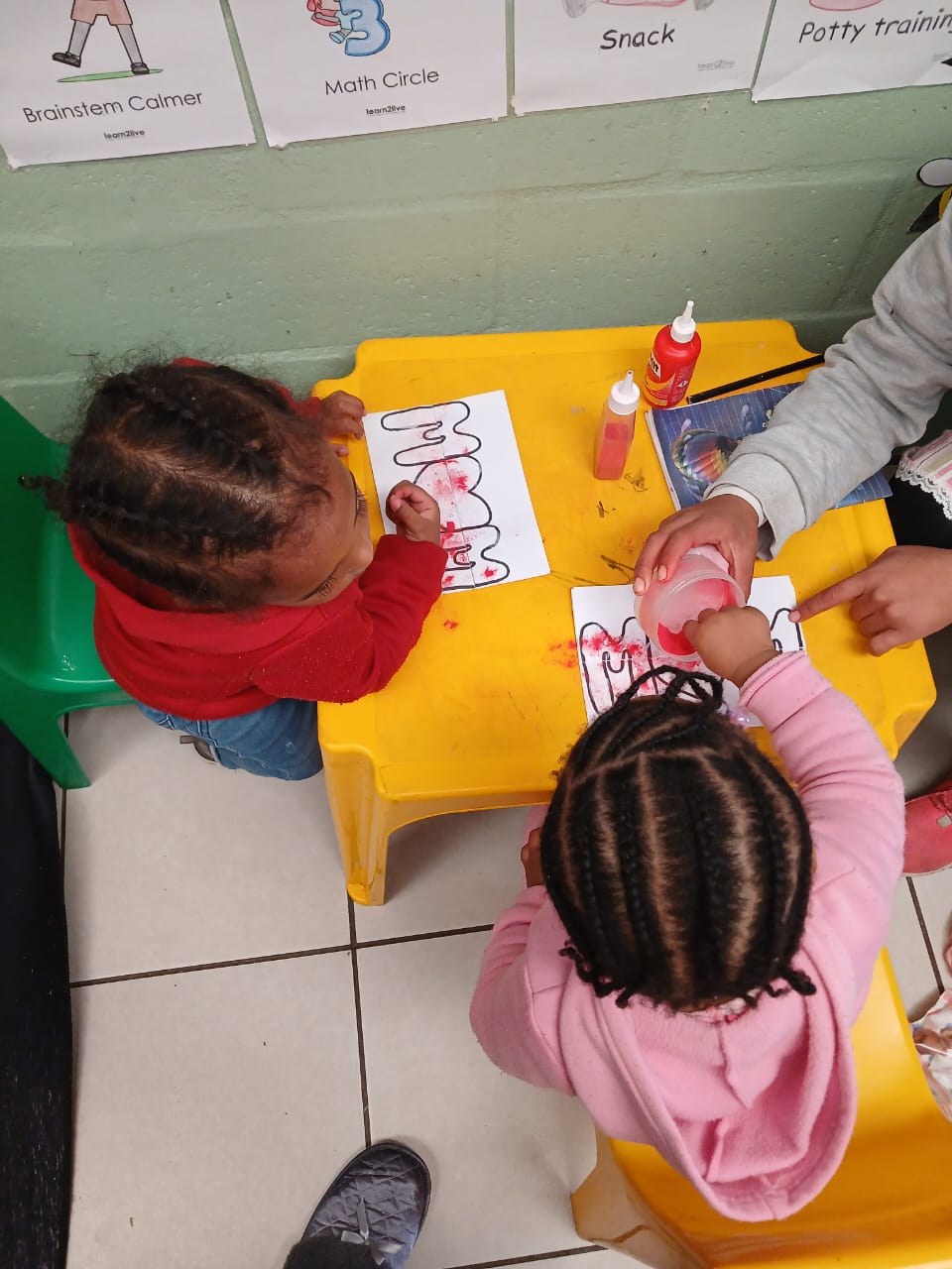 Young children engaged in creative art activities at a yellow table in a bright classroom with educational posters