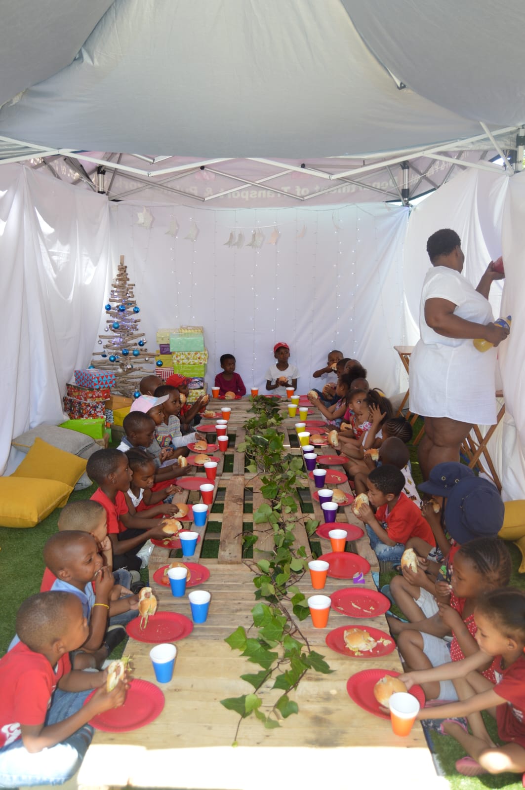 A group of children enjoying a festive meal in a white tent decorated with a Christmas tree, string lights, and green plants running down the center of a long wooden table