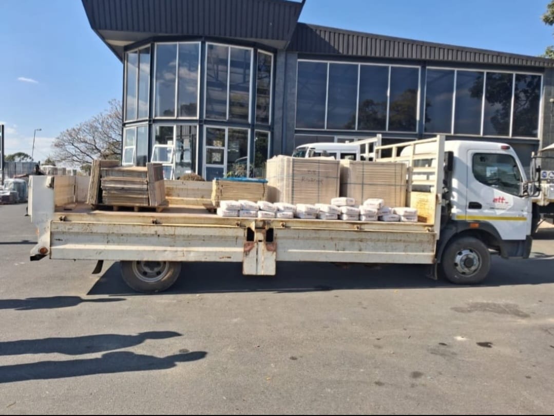 Delivery truck loaded with wooden fencing materials and construction supplies in front of a modern industrial building
