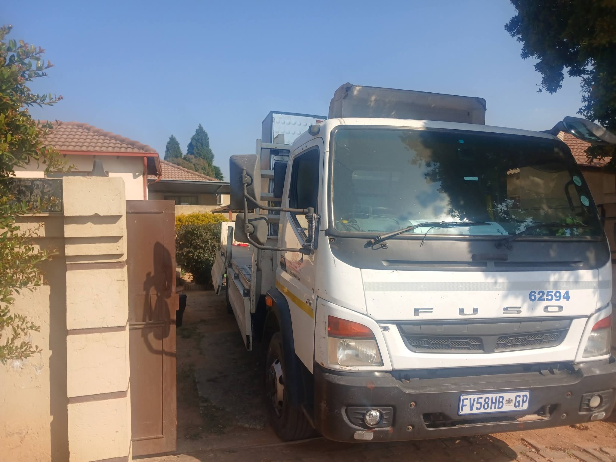 White Fuso delivery truck parked in a residential driveway next to a brown gate, with houses and trees in the background