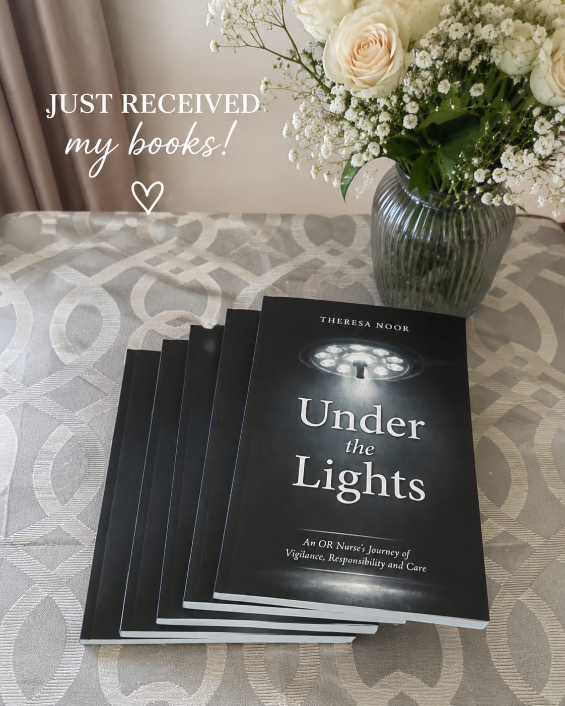 Stack of Under the Lights books by Theresa Noor displayed on a patterned bedspread next to cream roses and baby's breath flowers.