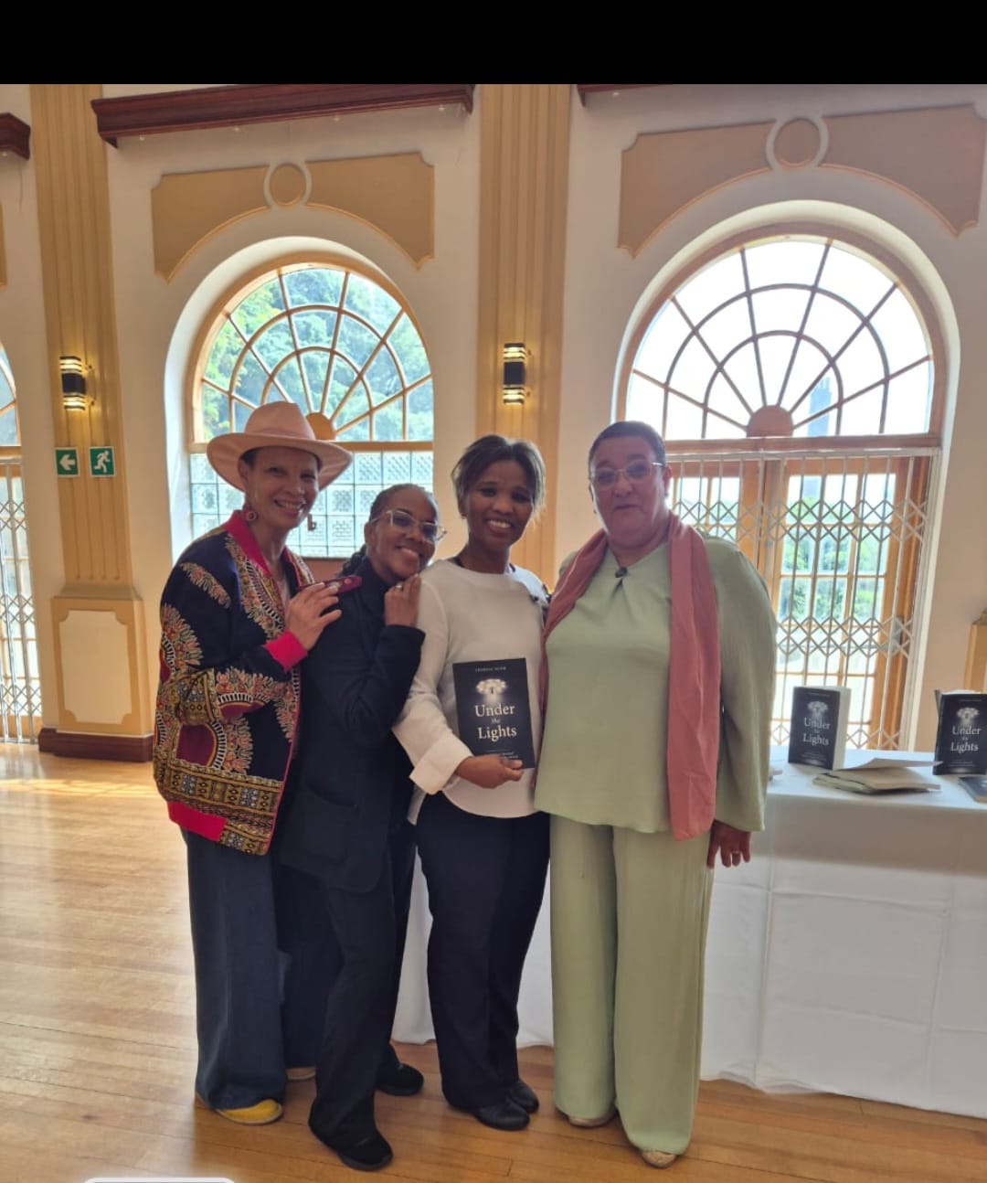 Four people pose together in an elegant venue with arched windows, with one person holding a book titled Under Lights.