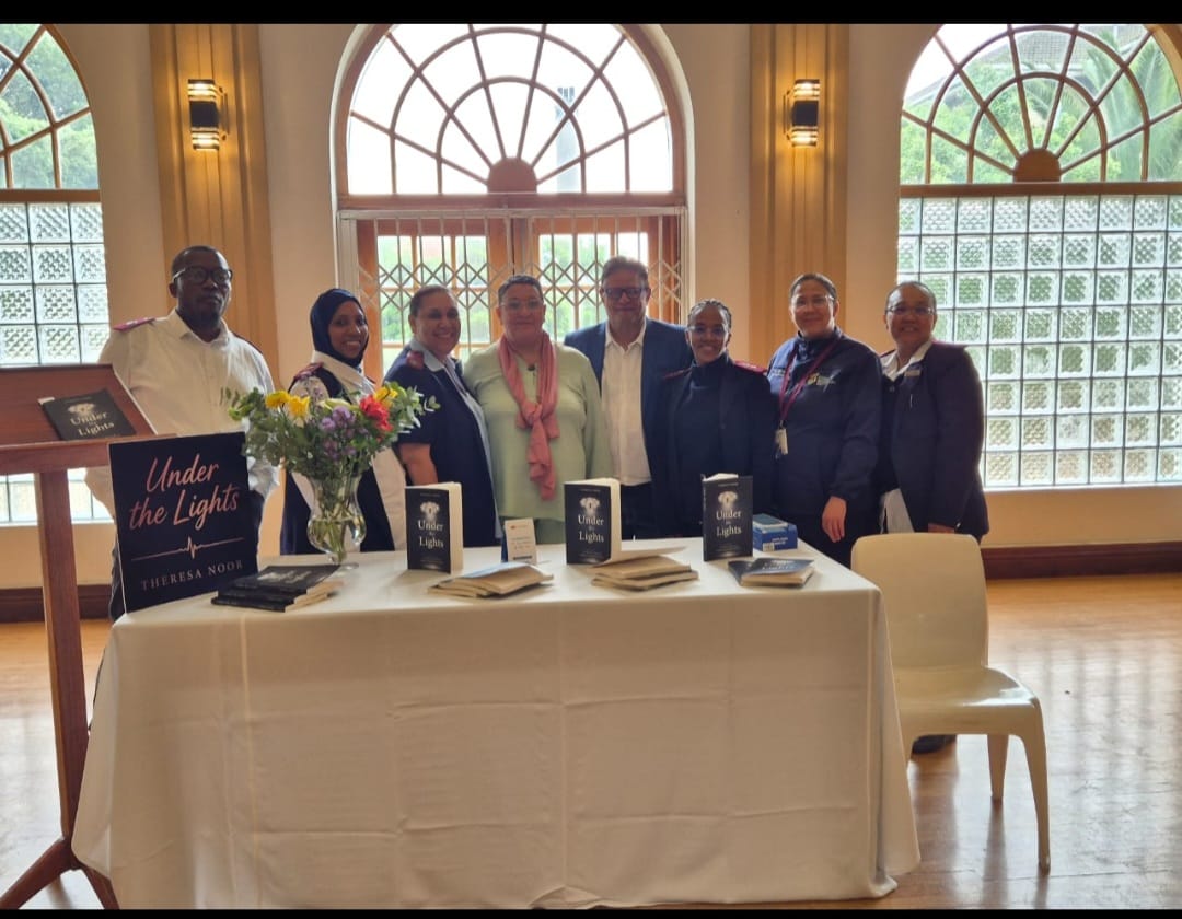 Group of eight people stand behind a white-draped table displaying books titled Under the Lights by Theresa Noor in an elegant venue with large arched windows and gold curtains.