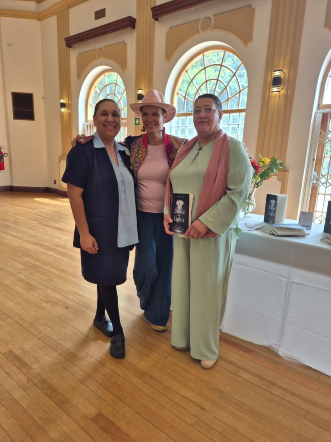 Three people pose together in an elegant hallway with arched windows, with the woman in the center holding an Under Lights award.