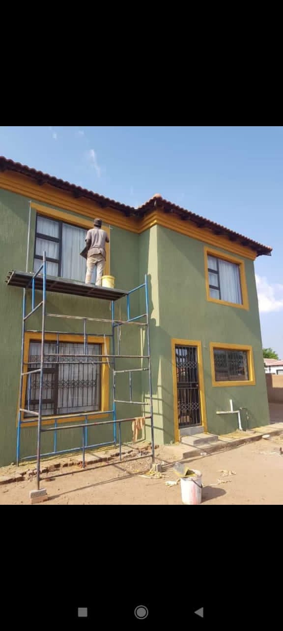 Worker on blue metal scaffolding painting the exterior of a two-storey house with green walls and yellow trim