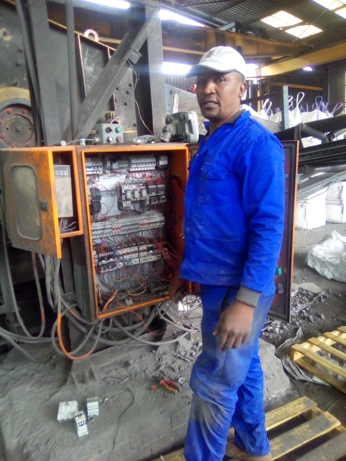 Peter Mashamaite in blue workwear and white hard hat next to an industrial electrical control panel