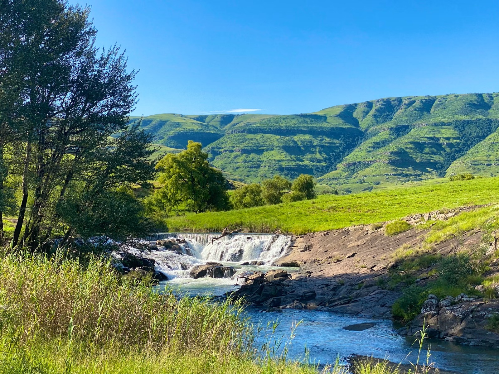 Sweeping mountain valley landscape with a flowing river and waterfall surrounded by lush green fields and verdant hills