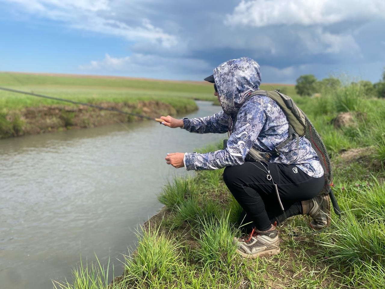 Person in protective gear crouching by a rural stream during environmental sampling